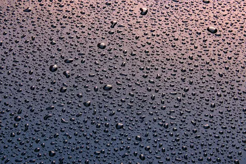 Numerous water droplets of varying sizes cover a smooth, dark surface, reflecting light