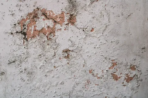 A close-up of a weathered concrete wall with peeling white paint and exposed pinkish-orange plaster underneath.